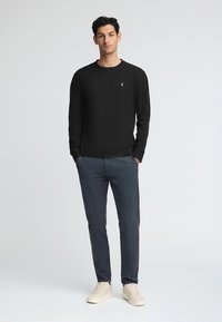 Svart långärmad tröja med rund halsringning och en liten logotyp, kombinerad med mörkgrå byxor och ljusbeige sneakers.