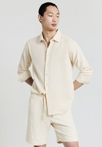 Uomo che indossa una camicia a maniche lunghe in crema testurizzata con bottoni e pantaloni corti abbinati, in piedi con le braccia dietro la schiena, mentre guarda di lato.