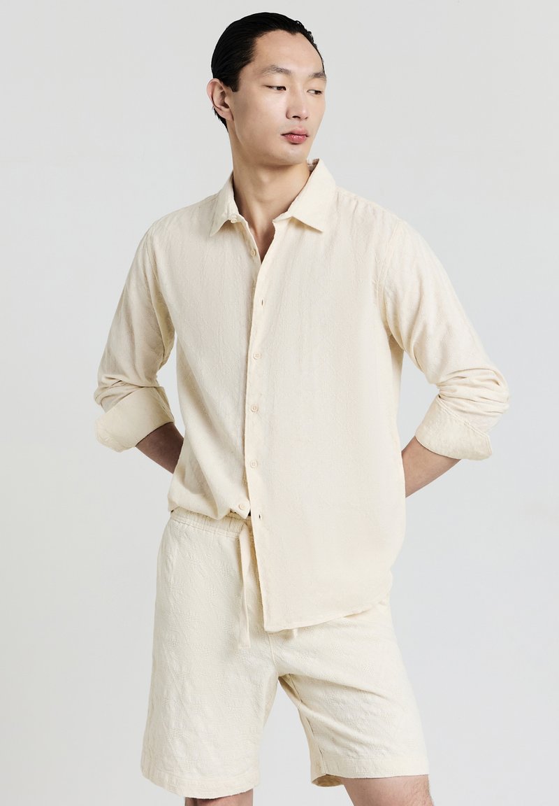 Uomo che indossa una camicia a maniche lunghe in crema testurizzata con bottoni e pantaloni corti abbinati, in piedi con le braccia dietro la schiena, mentre guarda di lato.