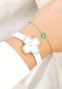 Montre avec un bracelet en cuir blanc et un cadran en forme de trèfle à quatre feuilles doré, visage blanc et accents dorés. Comprend un bracelet avec un charme en trèfle vert.