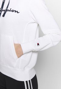 Felpa bianca con cappuccio caratterizzata da un logo "Champion" blu navy, una tasca frontale e una piccola toppa con logo sulla manica. Tessuto morbido con polsini a costine.