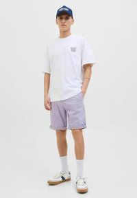 Witte katoenen t-shirt, lila shorts met opgerolde cuffs, witte sokken en sneakers met blauwe en beige accenten. Blauwe pet met logo.
