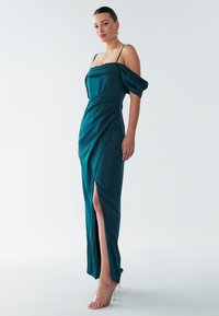 Teal satijnen lange jurk met off-shoulder mouwen, gestructureerd lijfje, gedrapeerde details en een hoge split. Doorzichtige bandjeshakken worden gedragen.