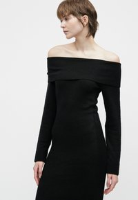 Kvinne iført en svart av-shoulder langermet tettsittende kjole, som ser til venstre mot en ensfarget hvit bakgrunn.