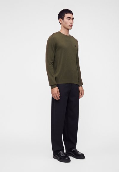 Homme debout, portant un pull à manches longues vert olive, un pantalon noir ample et des chaussures en cuir noires sur un fond blanc uni.