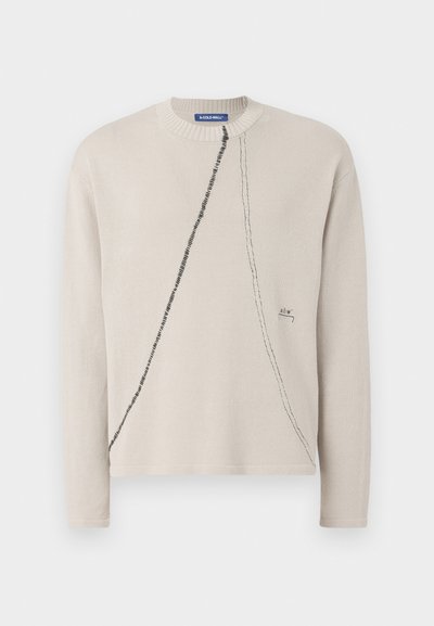 Bežový pletený sveter s okrúhlym výstrihom, ktorý má čierny diagonálny šev a vyšívané logo na leme. Mäkkučká, štruktúrovaná tkanina.