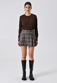 Cardigan marrone a maglia con bottoni, abbinato a una gonna pieghettata grigia a scacchi. Il modello indossa stivali neri alti fino al ginocchio e flat. Sfondo semplice.