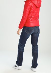 Rød pufferjakke med hætte, skinnende tekstur, parret med mørkeblå jeans. Hvide sneakers med sorte striber fuldender outfittet.