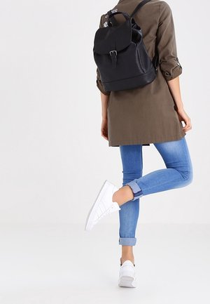 Black leather backpack with a flap and buckle detail, worn on a person in an olive green jacket and blue jeans, paired with white sneakers.