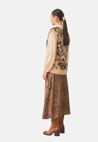 Chaleco de punto beige con patrones florales negros, superpuesto a una blusa de manga abullonada crema y una falda marrón con estampado. Botas con tacón marrones.