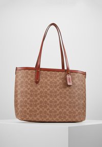 Sac fourre-tout marron à motifs avec des poignées en cuir et une petite étiquette en cuir suspendue sur un piédestal d'exposition blanc.