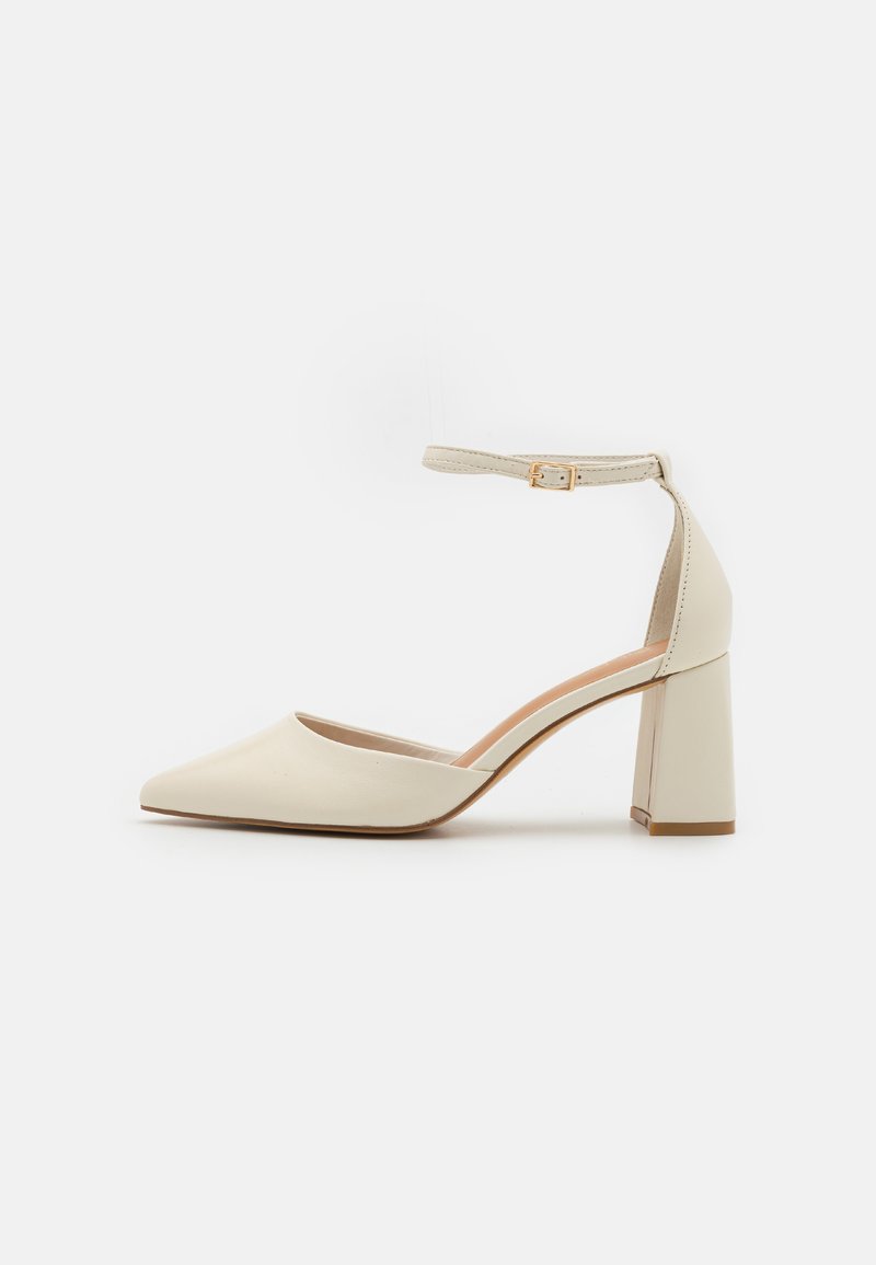 Décolleté in pelle beige con tacco blocco e punta appuntita, cinturino alla caviglia e texture liscia. Presenta un dettaglio in metallo dorato sulla fibbia del cinturino.