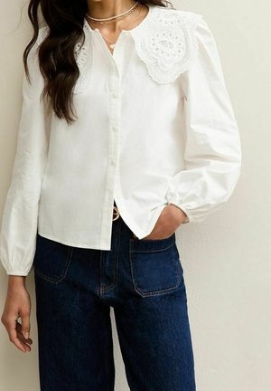 Biała bluzka z długim rękawem z dużym haftowanym koronkowym kołnierzem, w połączeniu z granatowymi, wysokim stanem dżinsami, dłoń w kieszeni.