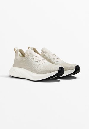 Light beige knit sneakers with thick white soles and black rubber toe grips, featuring slip-on design with laces and pull tabs.