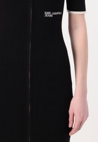 Juodas gulinys suknelė su priekiniais užtrauktukais, trumpomis rankovėmis su baltu apvadu ir balta siuvinėta "KARL LAGERFELD JEANS" logotipu.