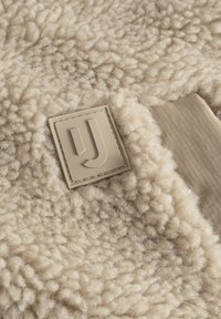 Beige stof med en fluffy, tekstureret overflade, der har en rektangulær logo-patch i en dæmpet tan farve. Et glat stofområde er synligt.