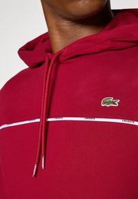Raudona kapučino su raišteliais, tekstūruota medžiaga, balta juostelė su "LACOSTE" prekės ženklu ir žalia siuvinėta krokodilo logo.