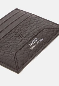 Pruunis tikitud nahast kaardihoidja, millel on kolm kaardipesa, kus on valget värvi väljatrükitud brändinimi "GUESS" ja "GENUINE LEATHER".