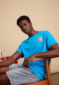 Blauwe katoenen T-shirt met korte mouwen en grafische print. Gemaakt met lichtdenim korte broeken, zittend op een houten stoel. Neutraal achtergrond.