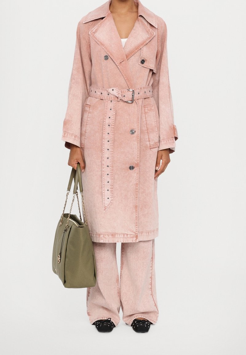 Personne portant un trench-coat rose ceinturé et un pantalon large assorti, tenant un grand sac à main vert olive avec des anses en chaîne, chaussures noires cloutées visibles.