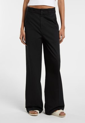 Person trägt schwarze, hoch taillierte, weit geschnittene Hose mit Knopf und Reißverschluss, kombiniert mit cremefarbenen Slip-on-Sandalen und einem kurzen weißen Top.