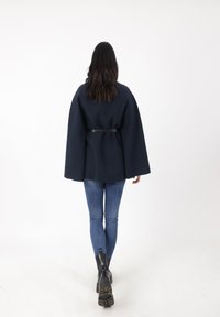 Capa navy con un diseño estructurado, ajustada a la cintura con un cinturón negro. Modelo vistiendo jeans azules ajustados y botines negros.