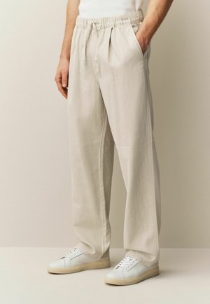 Person wearing beige drawstring pants with pockets and white sneakers, standing against a plain neutral background.