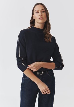 Maglione pullover blu navy con colletto alto e maniche a tre quarti, con piping crema lungo le maniche. Abbinato a jeans scuri.