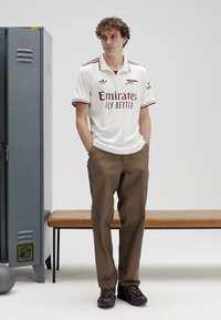 Jersey branco de manga curta com detalhes em vinho e texto, combinado com calças castanhas e sapatos escuros, contra um fundo neutro.