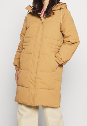 Kobieta ubrana w beżowy, puchowy płaszcz do kolan z kapturem, elastycznymi mankietami, przednimi kieszeniami i ukrytym zamkiem błyskawicznym, nosząca jasne spodnie.