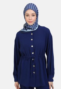 Marineblauwe blouse met knoopsluiting en een getailleerde taille, lange mouwen en goudkleurige knopen. Gepaard met een gemêleerde blauwe sjaal met witte accenten.