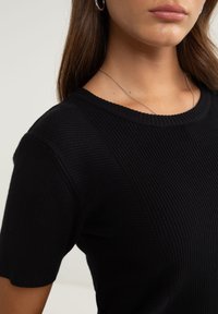Schwarzes, geripptes Strickoberteil mit kurzen Ärmeln und Rundhalsausschnitt, das eine texturierte Oberfläche und dezente Nahtdetails aufweist. Eine silberne Halskette ist sichtbar.