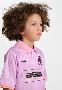 Rosa sporttröja med gula krageakcenter, med ett mönstrat mönster och logotyper för "hummel" och "BEES International."