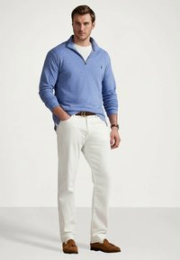 Man standing with one hand in pocket, wearing blue quarter-zip sweater, white pants, brown belt, watch, and brown tassel loafers.