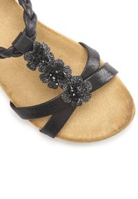 Schwarze Sandale mit geflochtenen Riemen und einem dekorativen Cluster aus schwarzen Perlen und Pailletten auf einer strukturierten, beigen Fußbett.