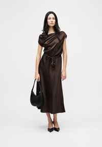 Γυναίκα με μακριά σκουρόχρωμα μαλλιά που φοράει midi φόρεμα από σατέν σκούρου καφέ χρώματος, δεμένο στη μέση, κρατώντας μια μαύρη τσάντα χειρός και φορώντας μαύρες μυτερές γόβες.