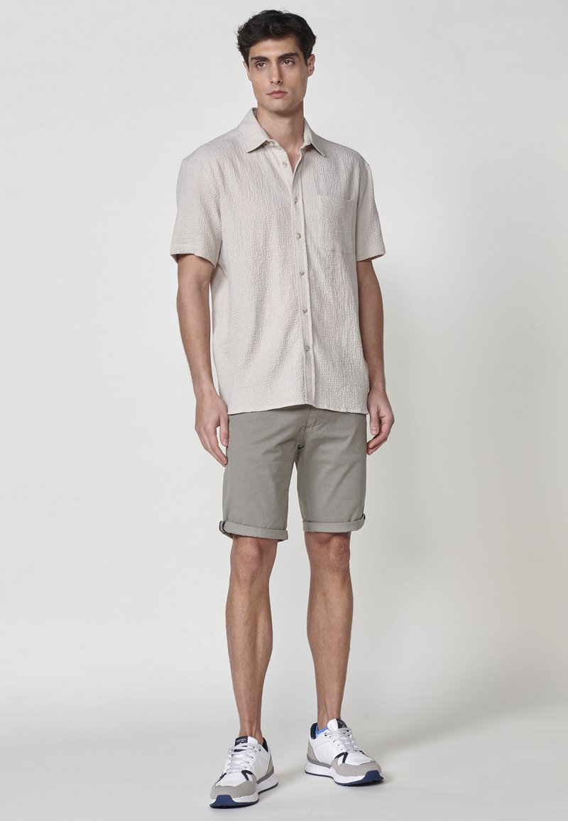Camicia beige a maniche corte con texture e taschino sul petto, abbinata a pantaloni corti verdi oliva arrotolati e sneakers bianche con dettagli navy.
