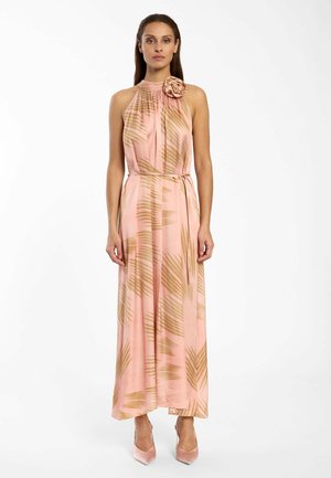 Frau steht und trägt ein ärmelloses pinkes Maxikleid mit braunen abstrakten Blattmustern, einem floralen Detail am Hals und pinken spitzen Absätzen.