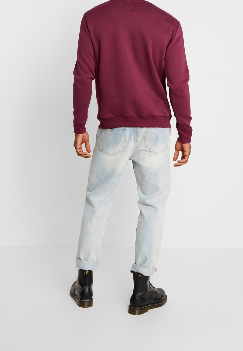 Homme portant une chemise à manches longues bordeaux, un jean bleu clair retroussé et des bottes noires à lacets, debout de dos sur fond blanc.