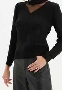 Maglione nero a coste con scollo a V e pannello in rete semi-trasparente, abbinato a pantaloni in pelle nera lucida; indossato con due anelli d'oro.
