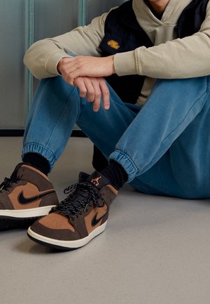 Person sitzt auf dem Boden und trägt braune und schwarze Nike Air Jordan Sneakers, blaue Jogginghosen, beige Kapuzenjacke und schwarze Weste.