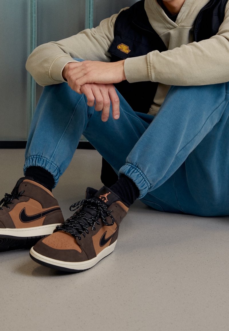 Personne assise par terre portant des baskets Nike Air Jordan marron et noires, un jogging bleu, un sweat à capuche beige et un gilet noir.