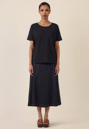 Woman with sleek hair wearing a black short-sleeve top, dark midi skirt, and brown platform sandals, standing against a plain background.