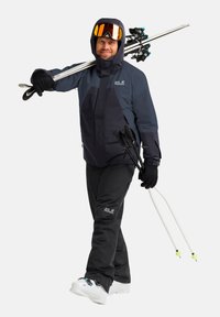 Abbigliamento da sci composto da una giacca impermeabile blu navy e nera, pantaloni neri isolati, guanti e occhiali da sci, mentre tiene dei bastoncini da sci.