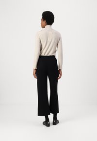 Bež pulover s turtleneck ovratnikom in dolgimi rokavi, v kombinaciji s črnimi širokimi hlačami. Model stoji z obrnjenim hrbtom proti kameri.