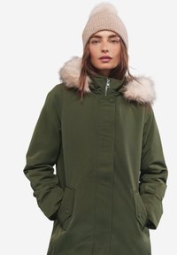 Parka verde oliva con cappuccio in pelliccia sintetica, zip frontale e tasche laterali. Accompagnata da un berretto in maglia beige chiaro con pompon sulla parte superiore.