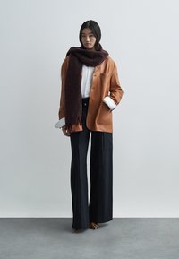 Brun läderjacka över vit skjorta, kombinerad med svarta byxor med vida ben och en mörk mönstrad halsduk. Fransar som detalj på halsduken.