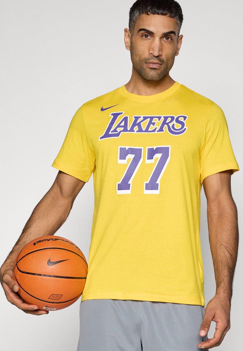 Gelbes T-Shirt mit kurzen Ärmeln, bedruckt mit "LAKERS" und der Nummer "77" in Lila, aus Baumwolle. Verfügt über einen Rundhalsausschnitt und wird mit grauen Shorts getragen.