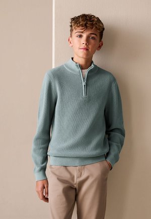 Una persona joven está apoyada contra una pared beige bicolor, vistiendo un suéter de punto azul claro con cremallera hasta un cuarto y pantalones beige.