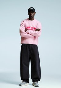 Moški stoji s prekriženimi rokami, oblečen v roza pulover z rdečim napisom, črne ohlapne hlače, sivo obuvalo in črno kapo z napisom "poskusi znova."